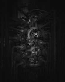 A complex network of industrial pipes and valves with a dark, grungy appearance. The metal surfaces are covered in rust and grime, indicating age and disuse.