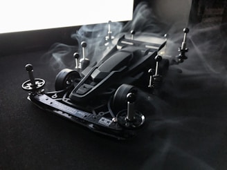 A sleek, futuristic model car is set on a dark surface with metal attachments and wheels. Surrounding the car, there is a misty smoke effect creating a mystical atmosphere. The image is lit from behind, casting shadows and highlighting the contours of the vehicle.