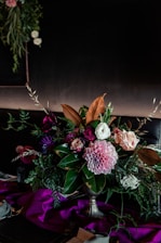 A lush and vibrant floral arrangement featuring a variety of flowers such as pink dahlias, roses, and ranunculus. The arrangement is surrounded by deep green foliage and sits in an elegant vase. The background is dark, creating a contrast that enhances the vivid colors of the flowers. A rich, purple fabric drapes across the bottom, adding a touch of luxury to the setting.