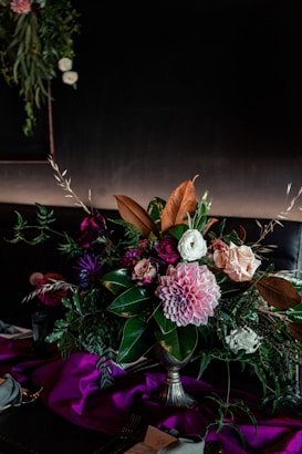 A lush and vibrant floral arrangement featuring a variety of flowers such as pink dahlias, roses, and ranunculus. The arrangement is surrounded by deep green foliage and sits in an elegant vase. The background is dark, creating a contrast that enhances the vivid colors of the flowers. A rich, purple fabric drapes across the bottom, adding a touch of luxury to the setting.
