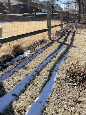 A wooden fence runs parallel to a patchy lawn with light snow covering some areas. Shadows from the fence slats create long, distinct patterns on the grass. In the background, there are houses and trees under a clear sky.