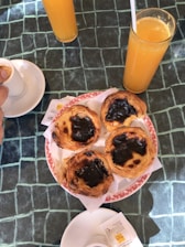 A plate of four pastries with a slightly charred surface is placed on a green checkered tablecloth. Accompanied by two glasses of orange juice and a white coffee cup with a hand holding it. Sugar packets are lying near the coffee cup.