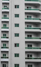 A modern residential building with multiple stories featuring uniform green-tinted glass balconies. The facade is painted in neutral white, and several windows are visible on each floor.