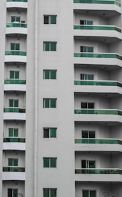 A modern residential building with multiple stories featuring uniform green-tinted glass balconies. The facade is painted in neutral white, and several windows are visible on each floor.