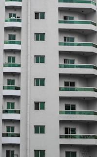 A modern residential building with multiple stories featuring uniform green-tinted glass balconies. The facade is painted in neutral white, and several windows are visible on each floor.