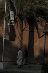 A tall palm tree casts a long shadow on a beige wall in the background, while two people walk by in the foreground. One person is dressed in a white traditional robe, and the other wears dark casual clothing. A large flag with Arabic text and graphic figures flaps in the breeze beside them.