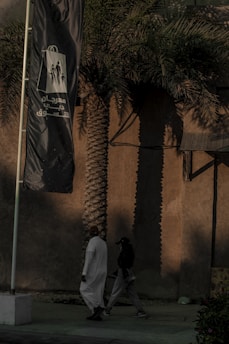 A tall palm tree casts a long shadow on a beige wall in the background, while two people walk by in the foreground. One person is dressed in a white traditional robe, and the other wears dark casual clothing. A large flag with Arabic text and graphic figures flaps in the breeze beside them.