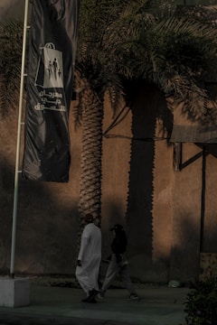 A tall palm tree casts a long shadow on a beige wall in the background, while two people walk by in the foreground. One person is dressed in a white traditional robe, and the other wears dark casual clothing. A large flag with Arabic text and graphic figures flaps in the breeze beside them.