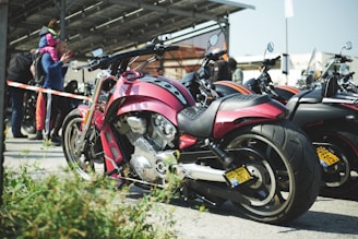 A red motorcycle is prominently parked among a row of other motorcycles. The setting appears to be an outdoor event, with people in the background gathered near a building with a canopy. A person is seen carrying a child on their shoulders, and another person seems to be speaking on a smartphone. The scene suggests a casual, social atmosphere.