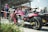 A red motorcycle is prominently parked among a row of other motorcycles. The setting appears to be an outdoor event, with people in the background gathered near a building with a canopy. A person is seen carrying a child on their shoulders, and another person seems to be speaking on a smartphone. The scene suggests a casual, social atmosphere.