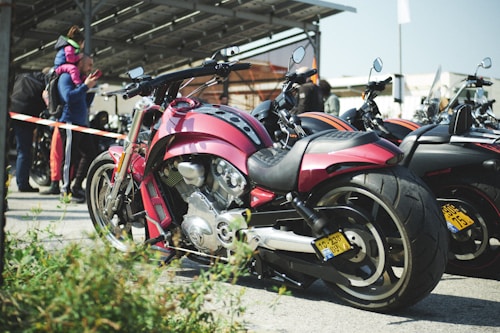 A red motorcycle is prominently parked among a row of other motorcycles. The setting appears to be an outdoor event, with people in the background gathered near a building with a canopy. A person is seen carrying a child on their shoulders, and another person seems to be speaking on a smartphone. The scene suggests a casual, social atmosphere.