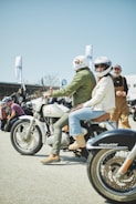 A group of people gathered outdoors for a motorcycle event. Two individuals wearing helmets are sitting on a vintage-style motorcycle, while others stand and converse in the background. The atmosphere is casual and lively, with people dressed in various styles of biker attire.
