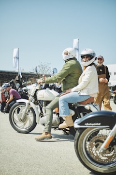 A group of people gathered outdoors for a motorcycle event. Two individuals wearing helmets are sitting on a vintage-style motorcycle, while others stand and converse in the background. The atmosphere is casual and lively, with people dressed in various styles of biker attire.