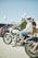 A group of people gathered outdoors for a motorcycle event. Two individuals wearing helmets are sitting on a vintage-style motorcycle, while others stand and converse in the background. The atmosphere is casual and lively, with people dressed in various styles of biker attire.