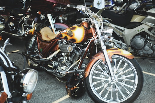 A custom motorcycle with a prominent flame paint job on the fuel tank and fenders is parked among other bikes. The chrome detailing and large front wheel give it an eye-catching appearance. The seat is tan leather, adding a vintage touch to the modern design.
