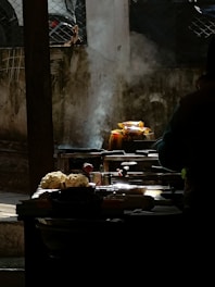 A street food stall setup is captured in low light with highlighted features. Smoke rises from cooking equipment, enhancing the scene's authenticity. Cauliflower heads and various utensils are displayed prominently, with a blurred figure suggesting activity.