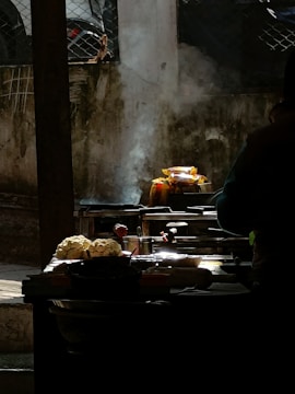 A street food stall setup is captured in low light with highlighted features. Smoke rises from cooking equipment, enhancing the scene's authenticity. Cauliflower heads and various utensils are displayed prominently, with a blurred figure suggesting activity.