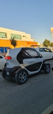 A small, compact electric car is driving on a street past a supermarket with orange signage. The car has a unique, futuristic design with a white and black exterior and blue-accented wheels. Other vehicles and trees are visible nearby, under a clear blue sky.