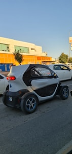 A small, compact electric car is driving on a street past a supermarket with orange signage. The car has a unique, futuristic design with a white and black exterior and blue-accented wheels. Other vehicles and trees are visible nearby, under a clear blue sky.