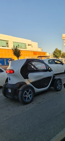 A small, compact electric car is driving on a street past a supermarket with orange signage. The car has a unique, futuristic design with a white and black exterior and blue-accented wheels. Other vehicles and trees are visible nearby, under a clear blue sky.