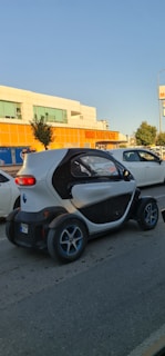 A small, compact electric car is driving on a street past a supermarket with orange signage. The car has a unique, futuristic design with a white and black exterior and blue-accented wheels. Other vehicles and trees are visible nearby, under a clear blue sky.