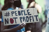 A person wearing a black mask holds a cardboard sign with the words '#PEOPLE NOT PROFIT' written in bold letters. The background is blurred, suggesting a public gathering or protest.