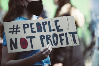 A person wearing a black mask holds a cardboard sign with the words '#PEOPLE NOT PROFIT' written in bold letters. The background is blurred, suggesting a public gathering or protest.