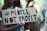 A person wearing a black mask holds a cardboard sign with the words '#PEOPLE NOT PROFIT' written in bold letters. The background is blurred, suggesting a public gathering or protest.