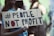 A person wearing a black mask holds a cardboard sign with the words '#PEOPLE NOT PROFIT' written in bold letters. The background is blurred, suggesting a public gathering or protest.