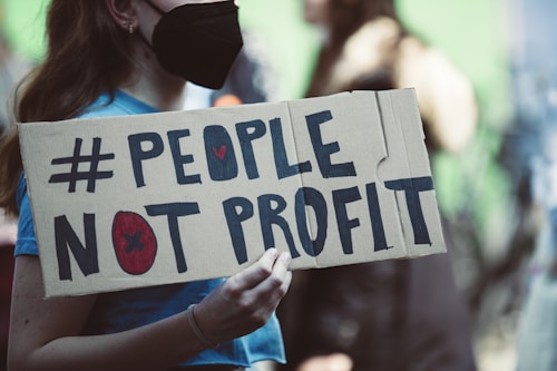 A person wearing a black mask holds a cardboard sign with the words '#PEOPLE NOT PROFIT' written in bold letters. The background is blurred, suggesting a public gathering or protest.