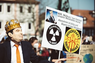A person in a suit and an orange tie wears a mask and a golden crown, holding a protest sign. The sign features multiple symbols and messages in German, referencing nuclear power and wind energy, with a satirical depiction of a public figure.