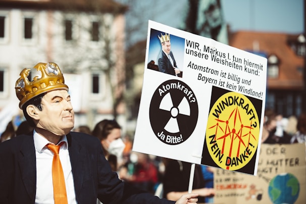 A person in a suit and an orange tie wears a mask and a golden crown, holding a protest sign. The sign features multiple symbols and messages in German, referencing nuclear power and wind energy, with a satirical depiction of a public figure.