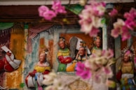 A wall mural depicting a group of traditional Asian figures dressed in colorful attire, surrounded by pink flowers in the foreground. The figures have detailed expressions and ornate headdresses, set against a multi-colored background with hints of architecture.