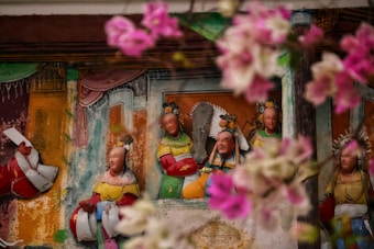 A wall mural depicting a group of traditional Asian figures dressed in colorful attire, surrounded by pink flowers in the foreground. The figures have detailed expressions and ornate headdresses, set against a multi-colored background with hints of architecture.