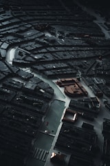 An overhead shot of a dark, intricate city model with winding streets and dense urban architecture, highlighted by sparse lighting that creates dramatic contrasts. The image conveys a sense of complexity and order within the labyrinthine arrangement of buildings and pathways.