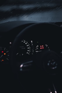 A dimly lit car dashboard with illuminated speedometer and warning lights. The background shows a rainy window, suggesting wet driving conditions.