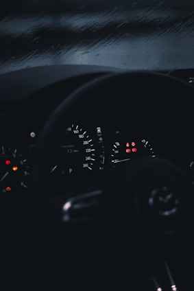 A dimly lit car dashboard with illuminated speedometer and warning lights. The background shows a rainy window, suggesting wet driving conditions.