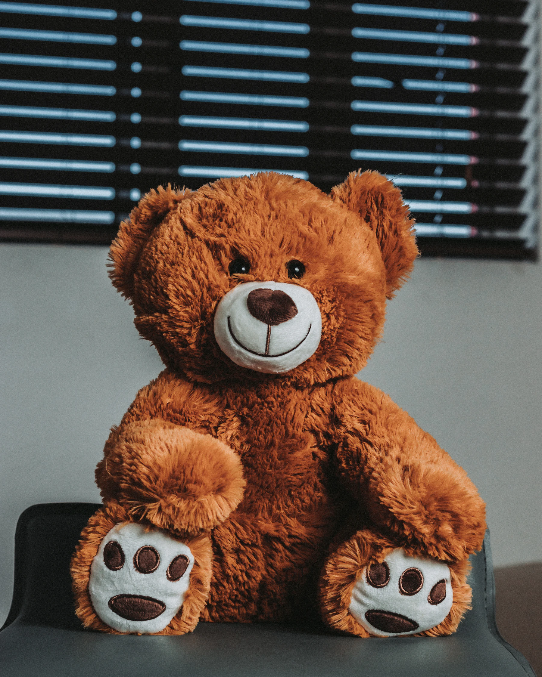 A brown teddy bear with white paw pads sitting on a gray chair in front of window blinds
