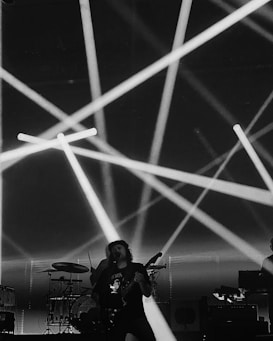 A black and white concert scene with dramatic lighting beams crisscrossing above a singer and musicians. The performer is playing a guitar, while a drum set and electronic equipment are visible in the background.