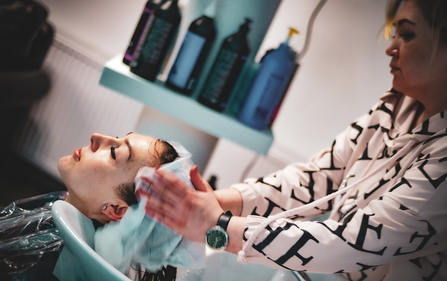 A man having his hair washed at the basin under a shelf of bottles