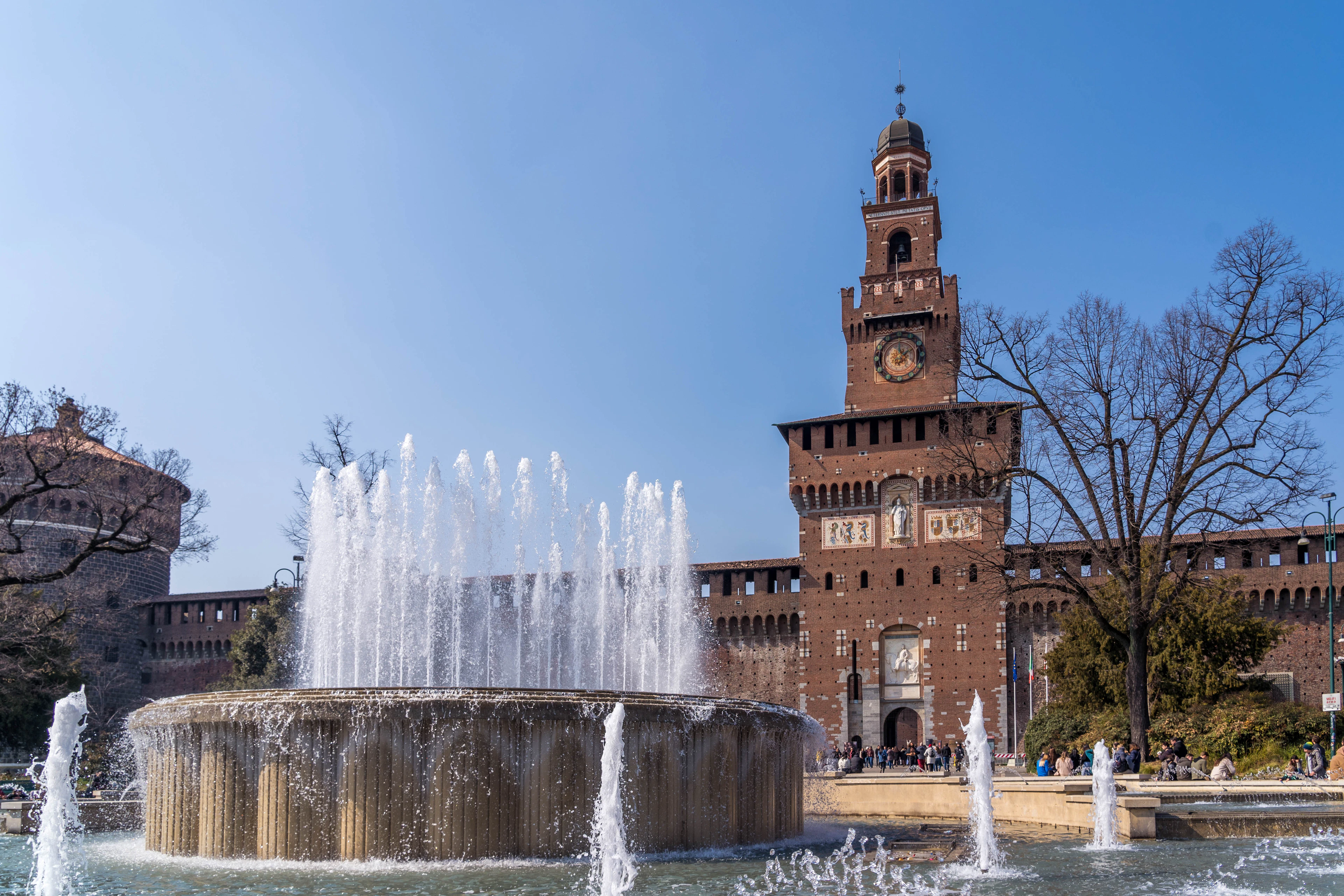 The great circular fountain in front of the Castello Sforzesco in Milan throwing up jets of water beneath the castle's red-brick clock tower