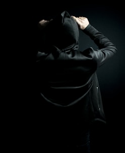 A person wearing a black hoodie with the hood up stands against a dark background. The focus is on the back and arms, which are raised, giving a sense of mystery and anonymity.