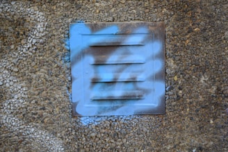 A metallic vent panel on a textured pebble-covered wall, partially covered with blue spray paint in a graffiti style.