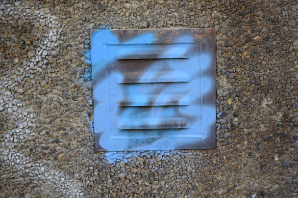 A metallic vent panel on a textured pebble-covered wall, partially covered with blue spray paint in a graffiti style.