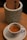 A small espresso cup filled with freshly brewed coffee sits on a white saucer. The cup features a logo with text, and in the background, there is a cylindrical, textured container holding what appears to be a menu or a cardboard item.
