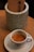 A small espresso cup filled with freshly brewed coffee sits on a white saucer. The cup features a logo with text, and in the background, there is a cylindrical, textured container holding what appears to be a menu or a cardboard item.