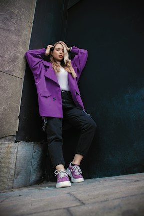 A person stands against a wall, striking a confident pose. They wear a fashionable purple jacket, black pants, and matching purple sneakers. The background is an urban setting with a mix of textured stone and dark-colored surfaces.