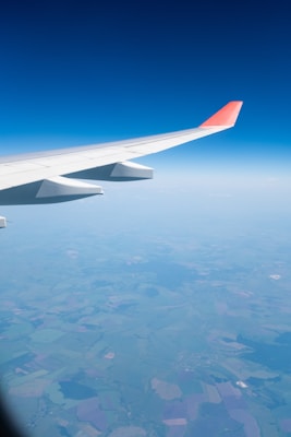 Aerial view of a landscape with sprawling fields under a blue sky. The aircraft's wing with an orange tip is prominently visible, casting a shadow over the earth below.