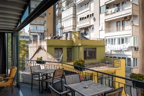 A balcony area with outdoor furniture including a table and chairs overlooks a cityscape of residential buildings. The buildings are multi-story with balconies, satellite dishes, and drying laundry. Several potted plants decorate the railing of the balcony.