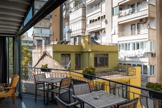 A balcony area with outdoor furniture including a table and chairs overlooks a cityscape of residential buildings. The buildings are multi-story with balconies, satellite dishes, and drying laundry. Several potted plants decorate the railing of the balcony.